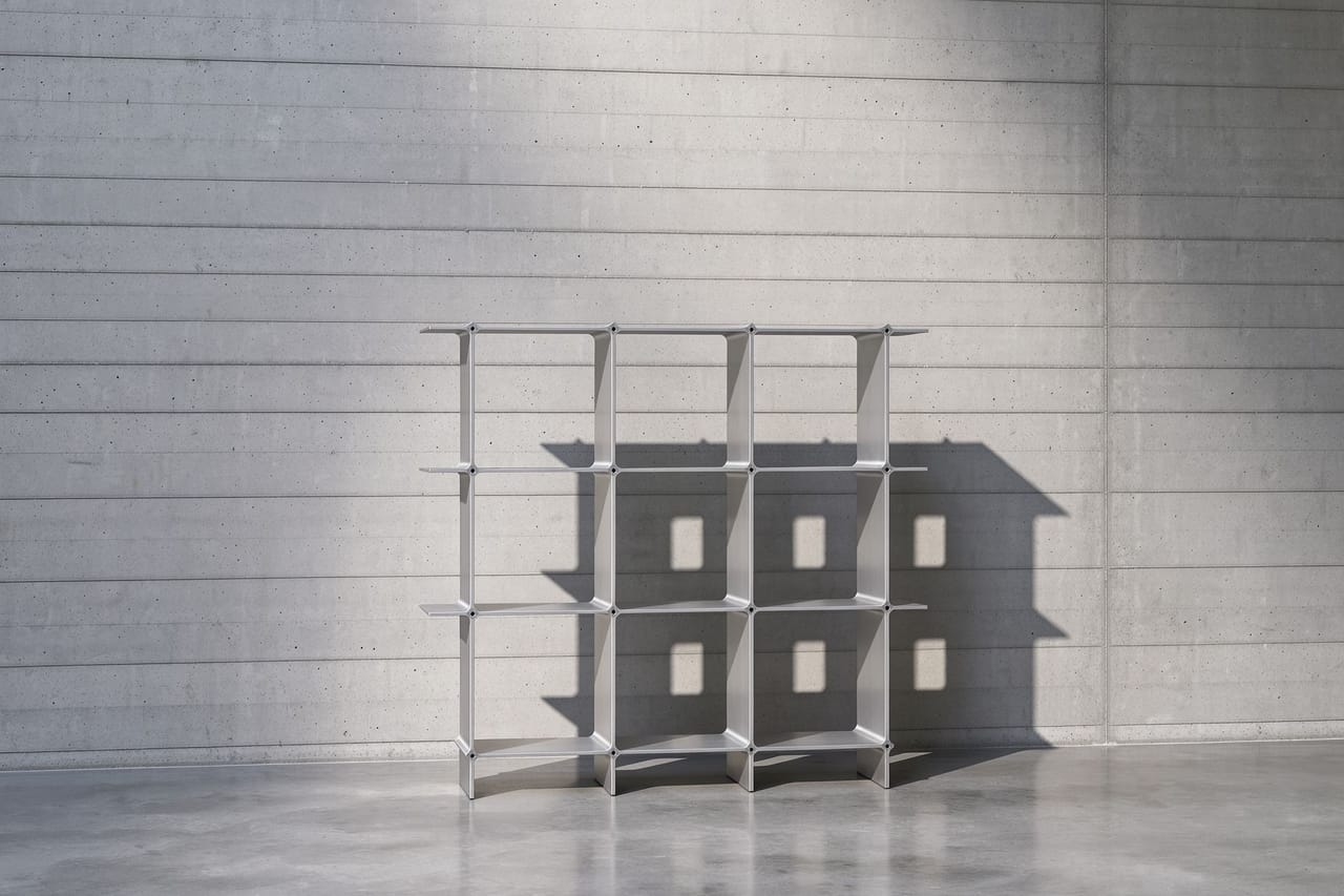 Montique cube shelf in silver, three by three cubes — in a hall of fair-faced concrete; the light falls from above and draws the open compartments as a shadow on the wall