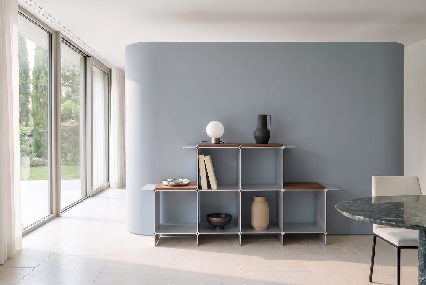 Montique shelving system in silver with wooden shelves in walnut, a globe lamp and a vase on top — against a curved blue-grey wall, on the left the glass front to the garden
